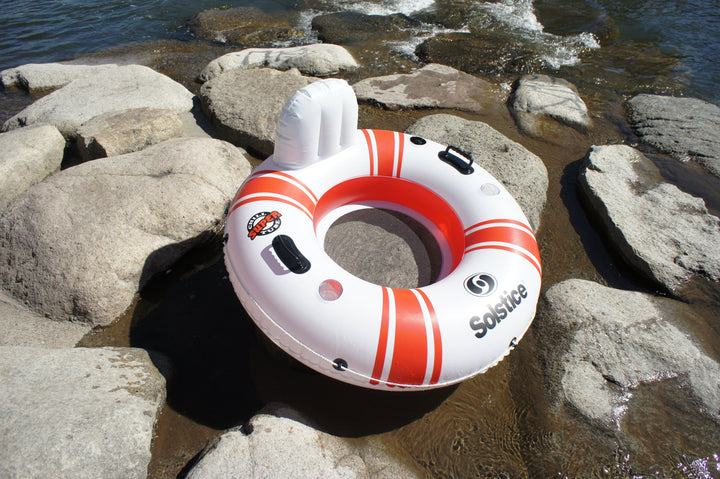 White and orange inflatable river tube with mesh seat and backrest lying on rocky riverbank near water