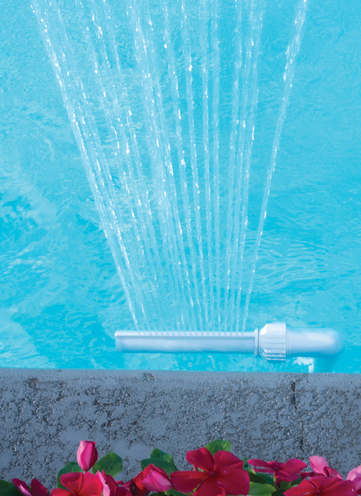 White water fountain spout spraying multiple streams of water into a bright blue pool near concrete edge with pink flowers