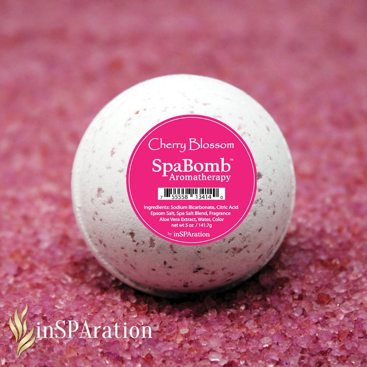 white spherical inSPAration SpaBomb Aromatherapy bath bomb with pink cherry blossom label sitting on coarse pink bath salts