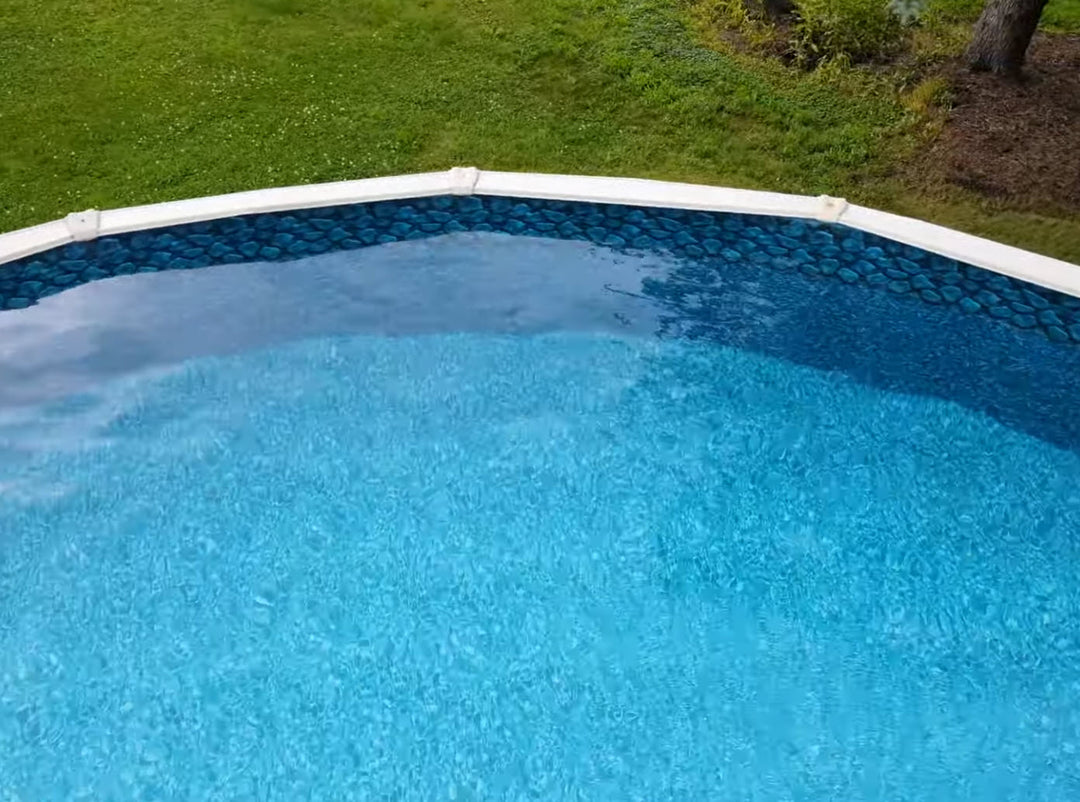 Top view of a round above ground pool with clear blue water and a decorative liner featuring a boulder and river rock pattern along the upper wall