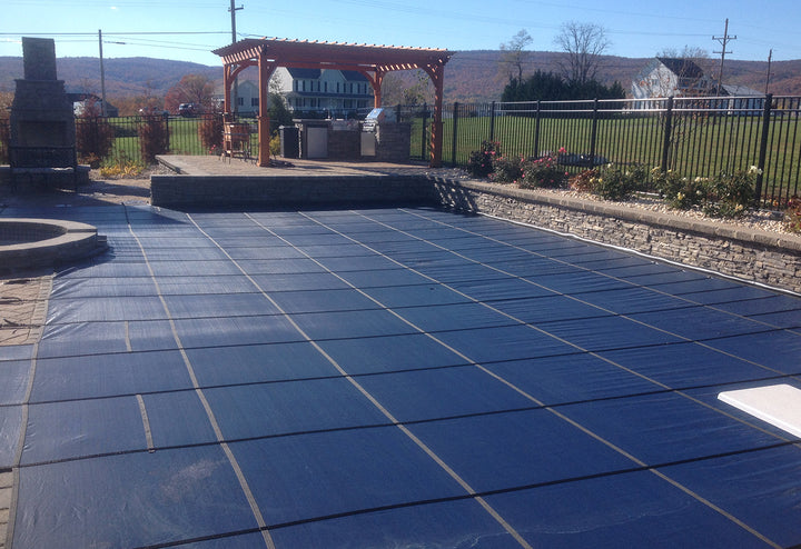 GLI 12 foot by 24 foot rectangle pool safety cover in blue mesh with black reinforcement straps secured over a rectangular in-ground pool