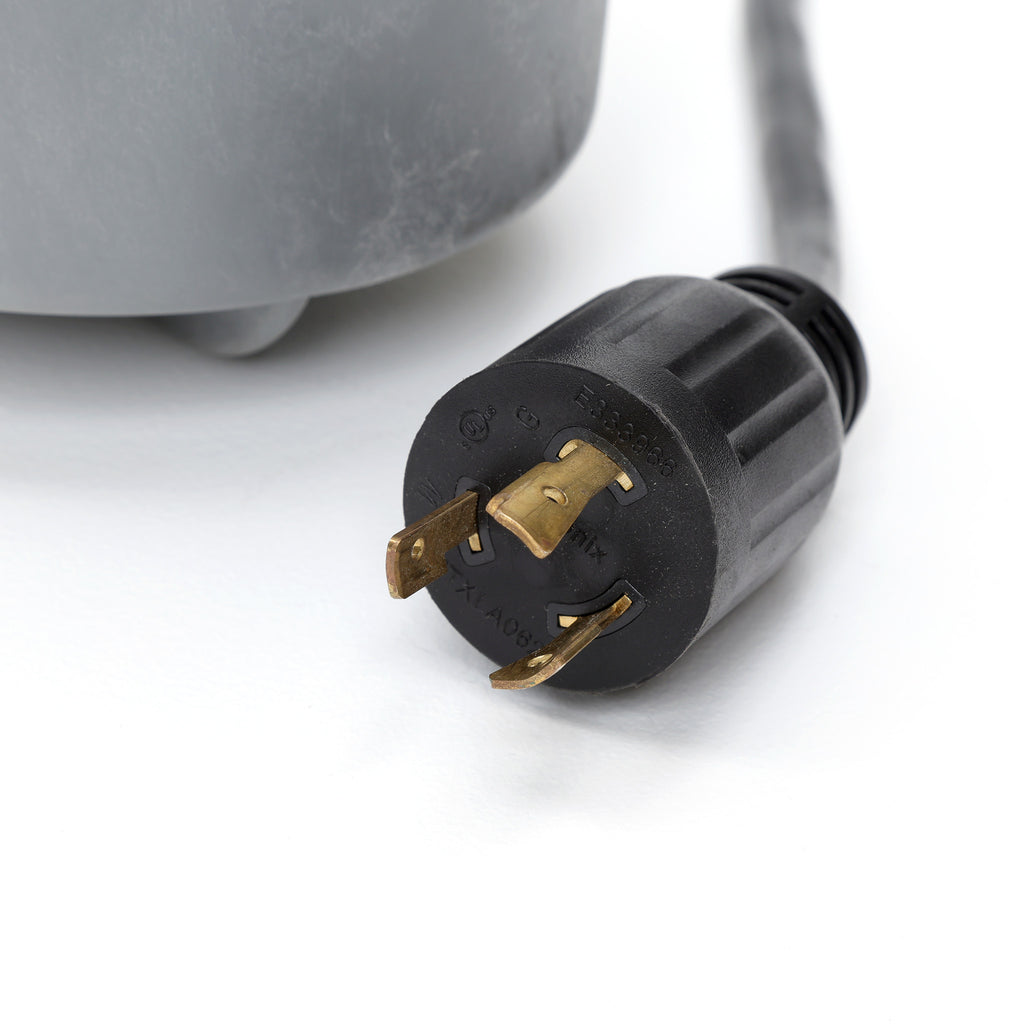 close-up on a black twist-lock electrical plug with three brass prongs attached to a gray water pump housing on a white surface