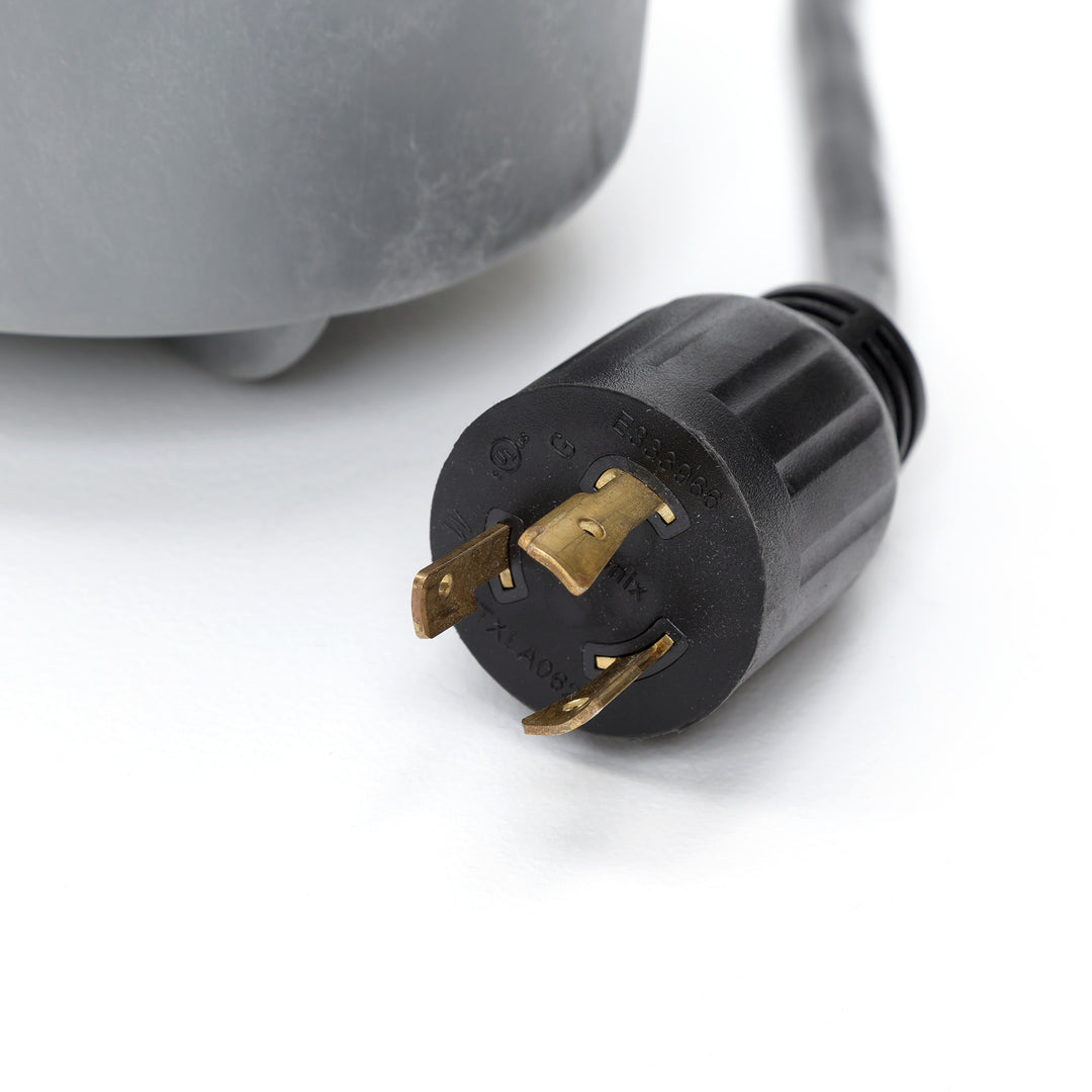 close-up on a black twist-lock electrical plug with three brass prongs attached to a gray water pump housing on a white surface