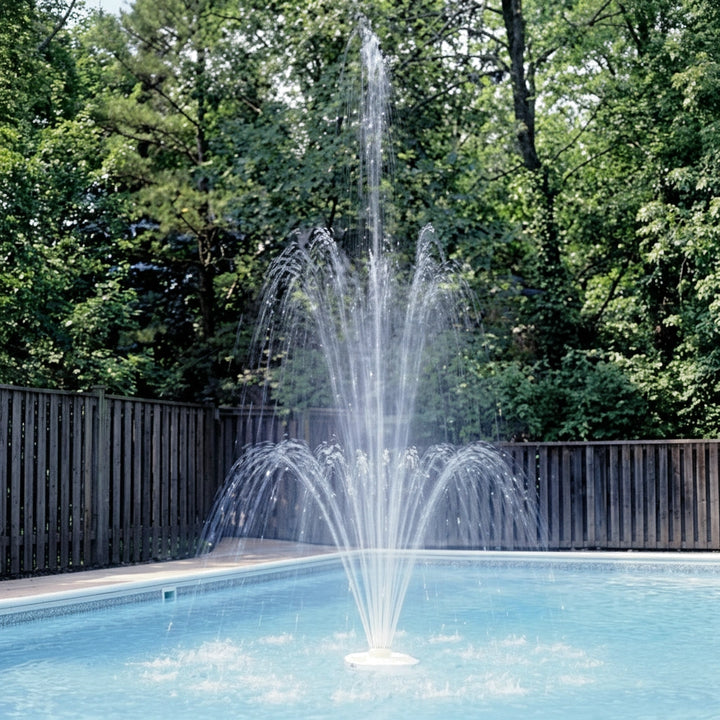A triple tier pool fountain spraying multiple arcs of water in a clear blue pool surrounded by a wooden fence and lush green trees